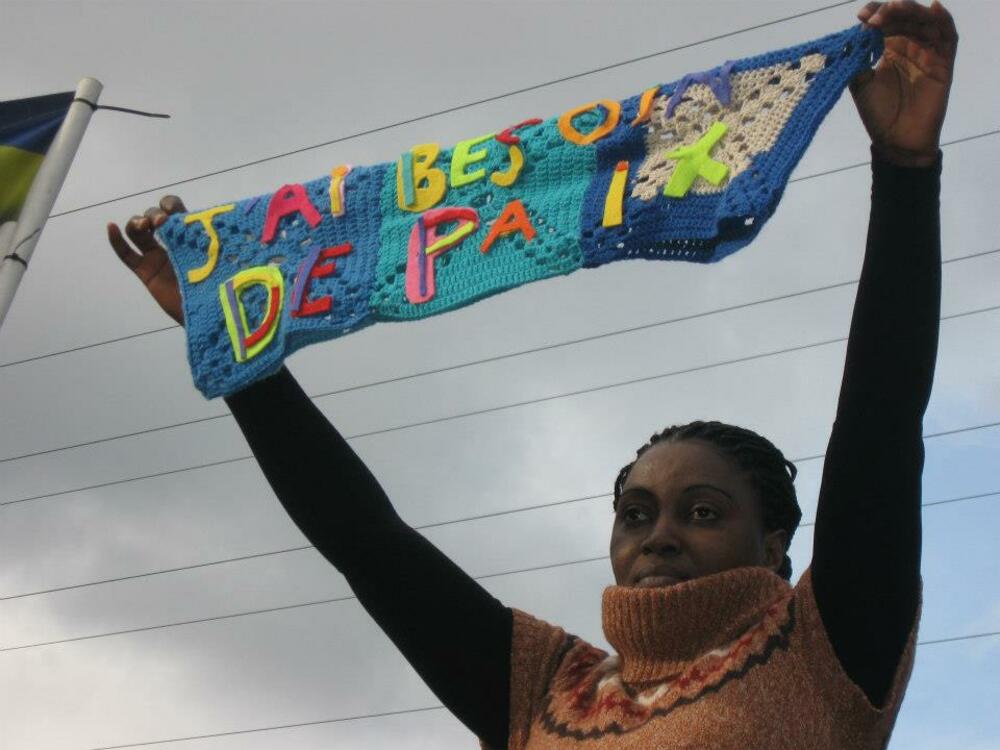 Une femme tenant une pancarte tricotée au-dessus de sa tête. Ça dit: J'ai besoin de paix