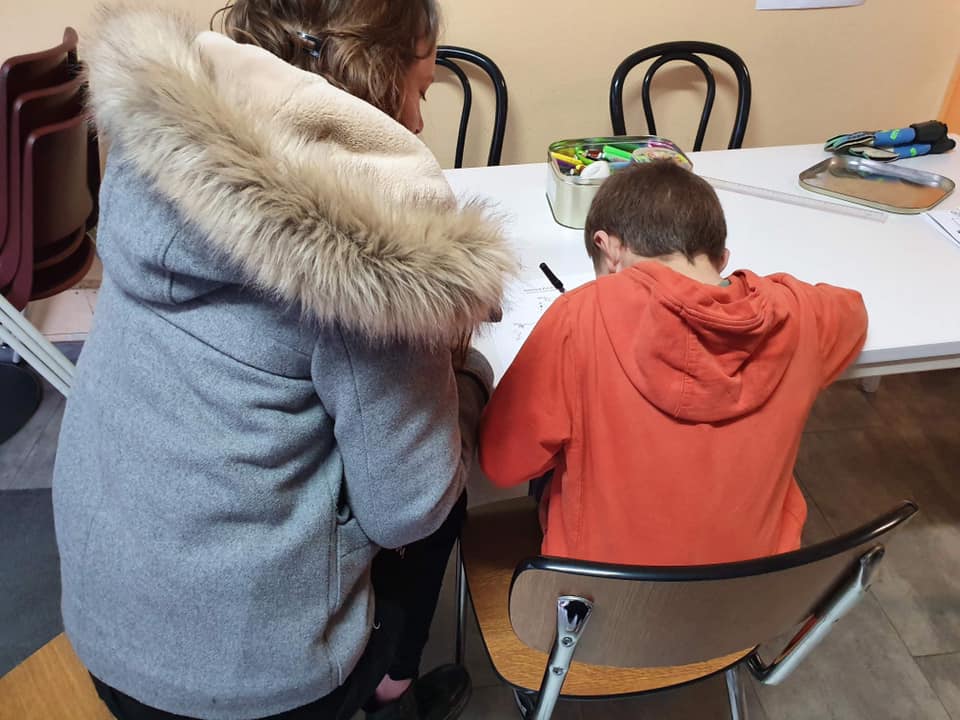 Une femme et un enfant assis à une table, dos à la caméra. L'enfant écrit dans un cahier. aide aux devoirs