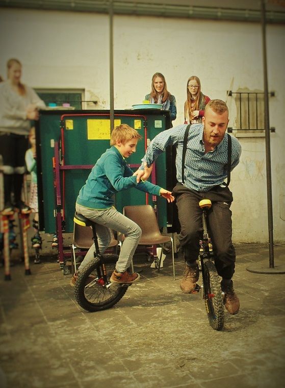 Un adulte et un enfant sur chacun leur monocycle, se tenant l'un à l'autre, roulant en cercle. Un groupe de jeunes adultes les regarde en arrière-plan. Tout le monde sourit
