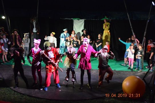 Beaucoup d'enfants de tous âges, déguisés et jouant, debout face à la caméra dans un cirque ou sur une scène