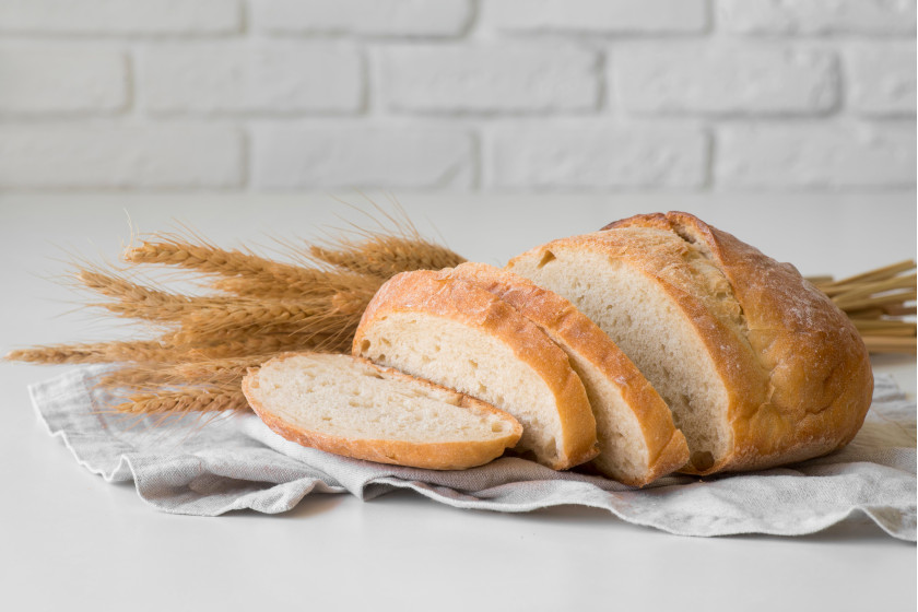 Une miche de pain, partiellement coupée en tranches. Derrière le pain se trouvent quelques tiges de céréales. Tout est sur une serviette de cuisine sur un comptoir blanc