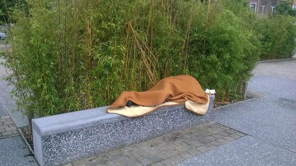 Sur un banc de pierre une personne enroulée dans une couverture.