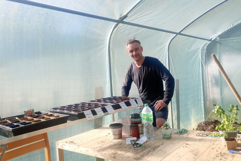 Dans une serre, un jeune homme devant une rangé de pots de plantes étiquetés.