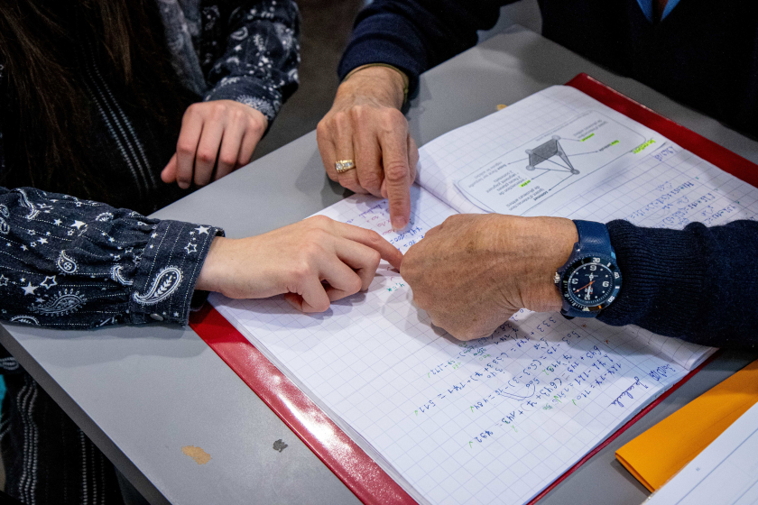 Deux paires de mains d’âges différents — un d’adulte plus âgé et un d’enfant — pointent ensemble un exercice dans un cahier ouvert rempli de notes et de schémas.